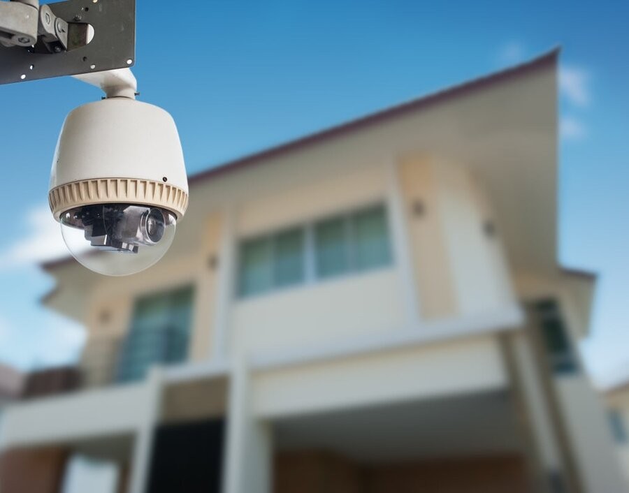 A smart surveillance camera in the foreground with a home’s exterior in the background.